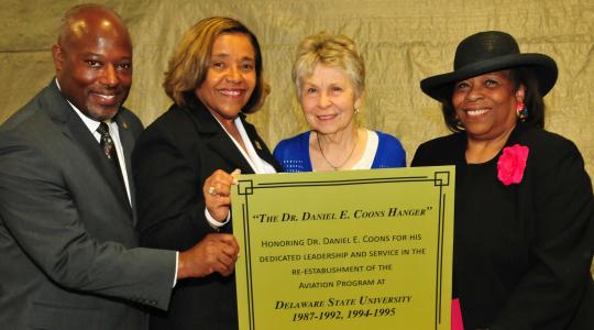 <p>(L-r) Lt. Col. Michael Hales, director of the DSU Aviation Program; College of Business Dean Donna Covington; JoAnn Coons, widow of Dr. Coons; Dr. Wilma Mishoe, DSU Board of Trustees member, hold a copy of the plaque that is being created for the Daniel E. Coons Hangar.</p>